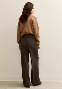 Maglione marrone con una vestibilità comoda, abbinato a pantaloni marroni a vita alta e a gamba larga, entrambi realizzati in tessuto morbido, su una superficie testurizzata.