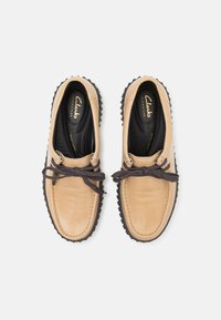 Beige skinnsko med rund tå og brune lisser, med en teksturert svart gummisåle og Clarks-merke inne.