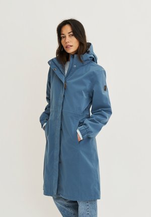 Vrouw draagt een lange blauwe regenjas met capuchon, handen in de zakken, staand tegen een effen lichte achtergrond, kijkt naar de camera.