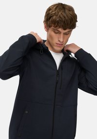 Marineblå softshell jakke med lynlås foran, justerbar hætte og sidelommer. Glat tekstur og minimalistisk design med diskrete stingdetaljer.