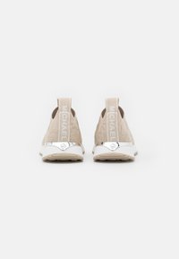 Para beżowych butów typu slip-on z fakturowaną tkaniną, białymi podeszwami i oznaczonymi pętlami z tyłu na białym tle.