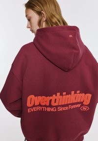 Sötétvörös, bő fazonú kapucnis pulóver kenguruzsebbel és nagy narancssárga hímzett felirattal a hátán: "Overthinking EVERYTHING Since Forever."