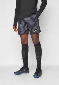 Svart langermet topp, mønstrede grå shorts med Nike-logo, svarte kompresjonsokker med hvite detaljer, og svarte fotballsko med blå detaljer.