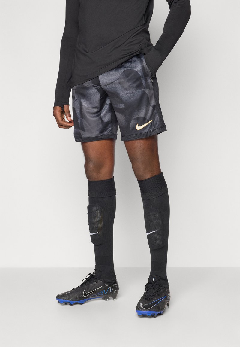 Svart langermet topp, mønstrede grå shorts med Nike-logo, svarte kompresjonsokker med hvite detaljer, og svarte fotballsko med blå detaljer.