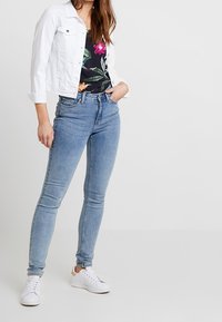 Giacca di denim bianca sopra una maglietta nera a fiori, abbinata a jeans skinny blu chiaro e sneakers bianche. La modella posa con una mano sul fianco.