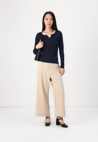 Marineblå langermet henley-overdel med knappdetalj, kombinert med vide beige bukser og svarte blokkhelsesko. Svart håndveske som tilbehør.