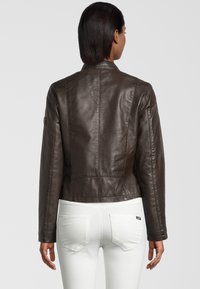 Bruine leren jas met een getailleerde pasvorm. Bevat een hoge kraag, naaddetails en een afgeronde zoom. Gedragen over een witte broek.