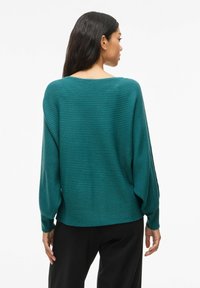 Pull en maille côtelée de couleur sarcelle avec une coupe ample, des épaules tombantes et des manches larges. Le dos présente un col arrondi et un motif texturé.