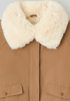 Veste couleur beige avec un col en fausse fourrure crème duveteuse, dotée de deux poches avant et d'une texture de tissu lisse. La fermeture à l'avant n'est pas visible.