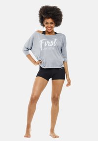 Gray off-shoulder long-sleeve top with white text “First I need coffee” paired with black shorts. Smooth fabric, relaxed fit, minimal design.