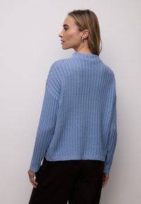 Pull en maille bleu clair avec un motif texturé, col montant et épaules tombantes ; porté avec un pantalon marron foncé.