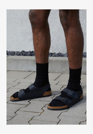 Černé sandály s korkovou stélkou, mají dvě nastavitelné spony, gumovou podrážku a měkký černý interiér, umístěné na dlažebním povrchu.