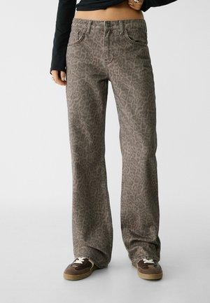 Vrouw draagt hoge, wijd uitlopende bruine jeans met luipaardprint, bruine sneakers en een zwart cropped top met lange mouwen.