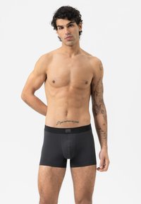 Schwarze Boxer-Briefs mit enganliegender Passform, aus glattem Stoff gefertigt. Sie verfügen über einen breiten Elastikbund mit einem kleinen Markenetikett. Das Modell steht mit entspannten Armen.