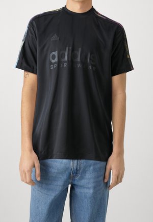 Person iført sort Adidas sportst-shirt med flerfarvede striber på ærmerne og lyseblå jeans, stående mod en ensfarvet baggrund.