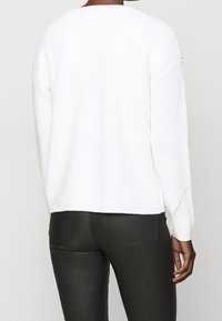 Persoon in een wit gebreid sweater en zwarte strakke broek, van achteren afgebeeld, staande tegen een effen achtergrond.