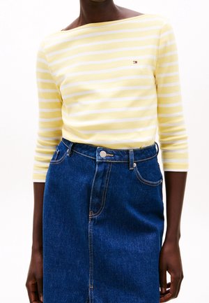 Persoon draagt een geel-wit gestreept shirt met lange mouwen, ingestopt in een blauw denim rokje met hoge taille en steekzakken aan de voorkant.