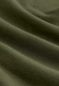 Weicher, dehnbarer Stoff in Olivgrün mit feiner Rippstruktur und diagonalen Falten, die eine glatte, gleichmäßige Oberfläche zeigen.