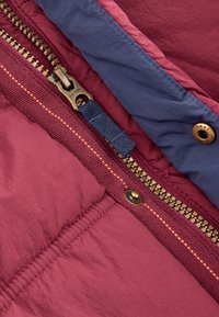 Veste bordeaux et marine avec une fermeture éclair visible, une quincaillerie de fermeture en bronze, des coutures contrastantes et une texture douce. Détail de la zone du col.