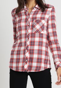 Camicia a maniche lunghe a quadri rossi e blu, realizzata in cotone, con taschino sul petto. Orlo leggermente curvo e design aderente.