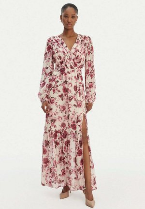 Donna in piedi che indossa un lungo vestito avvolgente a fiori con maniche lunghe e spacco laterale, abbinato a tacchi a punta beige su sfondo bianco.