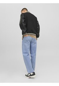 Svart varsityjacka med läderärmar, beige topp, ljusblå lösa jeans och svarta sneakers med vita detaljer, sedd bakifrån.