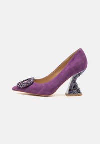 Tacco alto in suede viola con punta affusolata e dettaglio floreale decorato. Il tacco scultoreo presenta gemme viola e un design triangolare.