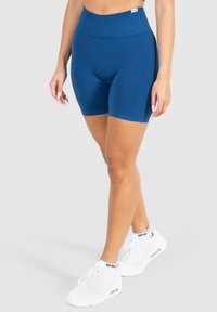 Blå atletiske shorts lavet af strækbart stof med en høj talje, der har en glat tekstur og en tætsiddende pasform, parret med hvide sneakers.