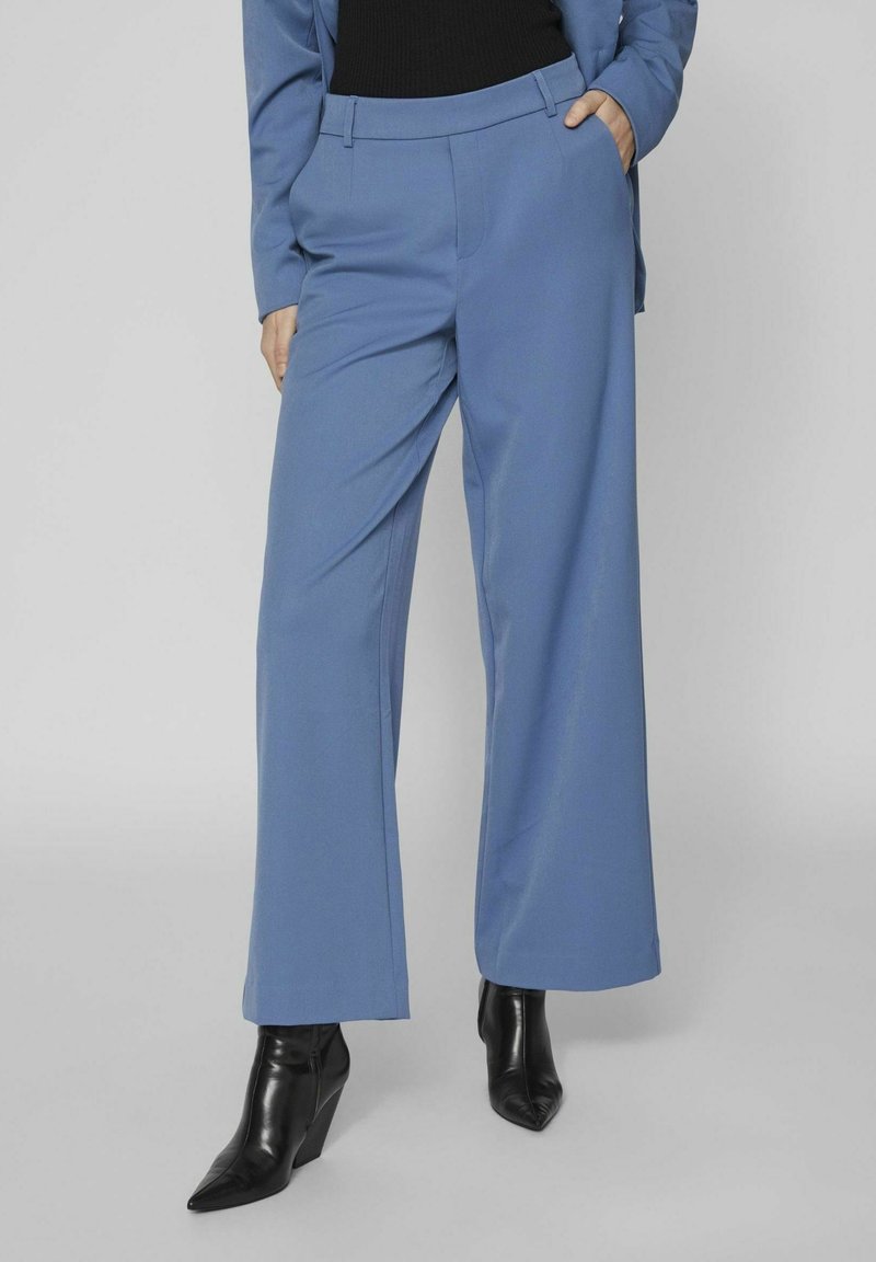 Femme portant un pantalon large bleu, un haut noir et des bottines noires à bout pointu, une main dans la poche, posant devant un fond uni.