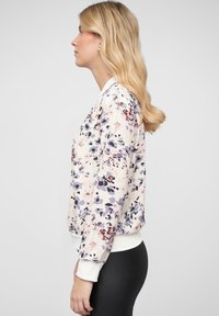 Bomber jacket dengan cetakan bunga yang menampilkan kain ringan, leher bulat, serta manset dan hem berwarna putih, dengan aksen ungu, pink, dan hitam.