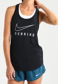 Canottiera nera da atletica con testo bianco "RUNNING" e logo Swoosh Nike. Indossata sopra uno strato bianco, abbinata a pantaloni corti turchesi. Texture liscia.