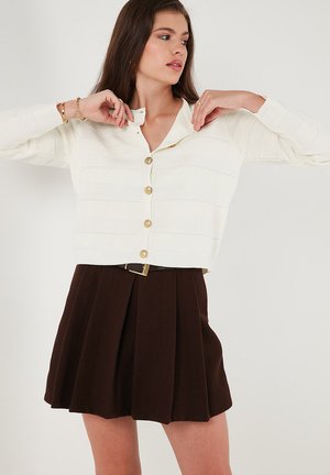 White, textured, long-sleeve cardigan with gold buttons, paired with a brown pleated skirt featuring a belt accent.