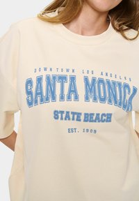 Krémové oversized tričko s modrým grafickým nápisem "SANTA MONICA STATE BEACH" a "DOWNTOWN LOS ANGELES", s kulatým výstřihem.