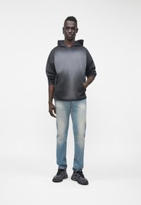 Νεαρός άνδρας στέκεται φορώντας ένα μαύρο και γκρι φούτερ με διαβάθμιση χρωμάτων, ξεθωριασμένο ανοιχτό μπλε τζιν και μαύρα αθλητικά παπούτσια, σε ένα απλό λευκό φόντο.