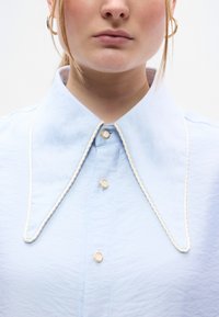 Ανοιχτό μπλε πουκάμισο με μεγάλος γιακά, με λευκή περίγραμμα στις άκρες και χρυσά κουμπιά. Λεπτή υφή υ φάσματος.