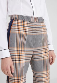 Pantalons à carreaux gris, orange et noir avec une taille élastique et des rayures contrastantes sur les côtés. Le matériau a une texture lisse.