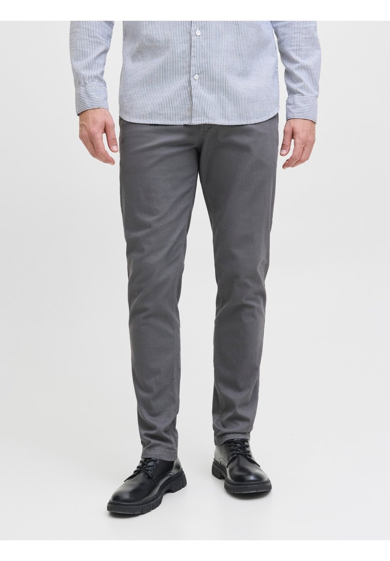 Homme portant un pantalon slim gris, des chaussures en cuir noires à lacets, et une chemise rayée bleu clair, debout devant un fond blanc.