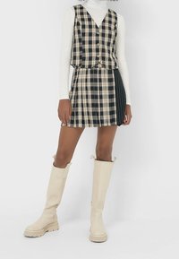 Robe à carreaux en noir, blanc et beige, dotée d'un haut de veste boutonné et d'une jupe plissée. Associée à des bottes beiges jusqu'au genou.