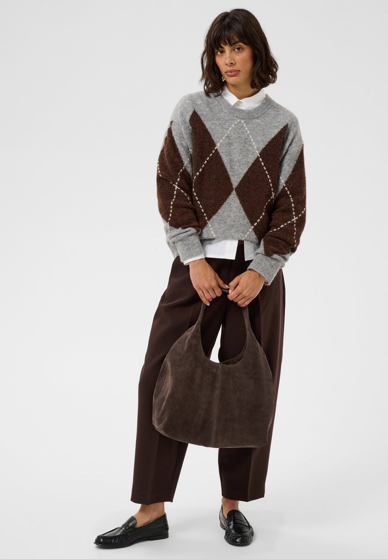 Femme portant un pull argyle gris et marron, un pantalon marron foncé, des mocassins noirs, tenant un grand sac à main en daim marron.