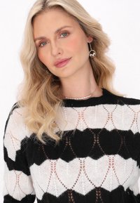 Maglione nero e bianco a maglia con un motivo a rosoni, caratterizzato da un design trasparente e testurizzato. Il modello indossa dettagli di perle e onde morbide.