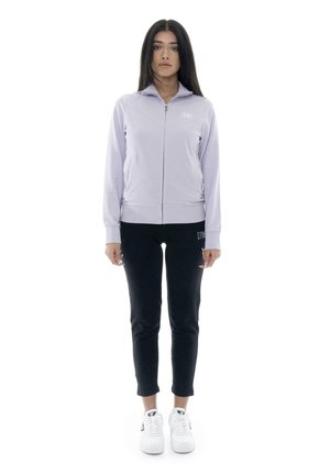 Une femme porte une veste zippée couleur lavande claire, un pantalon noir et des baskets blanches sur un fond blanc uni.