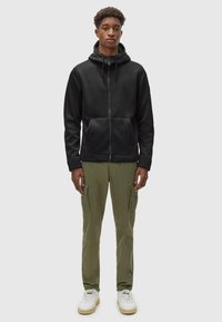 Felpa nera con zip e cordini, tasche frontali; abbinata a pantaloni cargo verde oliva, sneaker bianche con dettagli verdi.