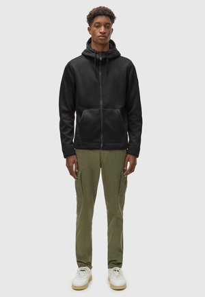 Black zip-up hoodie with drawstrings, front pockets; paired with olive green cargo pants, white sneakers with green accents.
