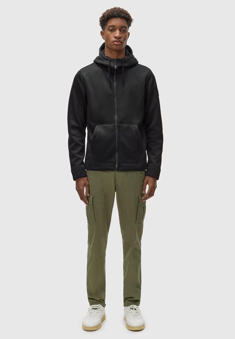Felpa nera con zip e cordini, tasche frontali; abbinata a pantaloni cargo verde oliva, sneaker bianche con dettagli verdi.