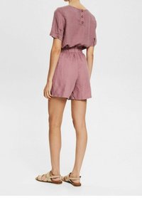 Combinaison en lin rose pâle avec manches courtes, taille élastique et détails à boutons au dos. Associée à des sandales à lanières beige clair.