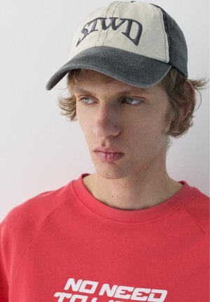 Mladi muškarac s valovitom plavom kosom nosi crvenu majicu dugih rukava sa natpisom "NO NEED" i bež-sivu kapu s izvezenim slovima.