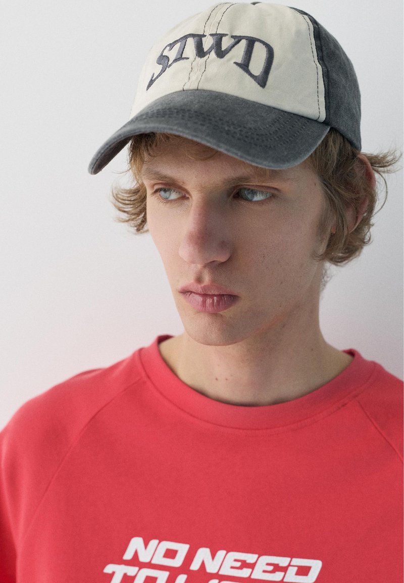 Jeune homme aux cheveux blonds ondulés porte un sweat-shirt rouge avec le texte "NO NEED" et une casquette beige et grise avec des lettres brodées.