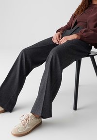 Personne assise sur une chaise noire portant un pantalon large gris foncé, une veste marron et des baskets beiges avec des lacets roses sur un sol blanc.