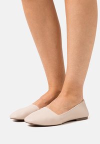 Ballerines beiges en matériau synthétique texturé avec un bout pointu et un talon bas et plat, présentant un design minimaliste et des lignes épurées.