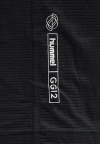 Czarna teksturowana tkanina z pionowym szwem, na którym znajduje się białe logo "hummel", geometryczny symbol powyżej oraz tekst "GG12" zamknięty w prostokącie.